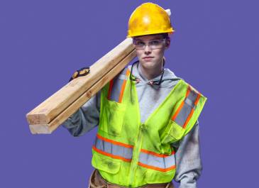 Woman wearing hard hat, safety glasses, gloves, hi-vis vest and holding two 2x4s over her shoulder.