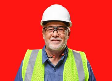 Man with white scruff wearing white hard hat, safety glasses, hi-visibility vest, and earplugs around his neck smiles at camera.