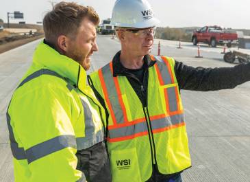 Two men wearing high visibility clothing and one wearing a WSI hard hat at road construction site.