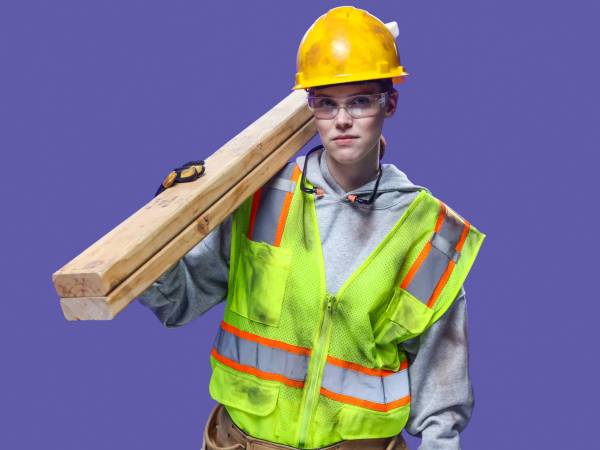 Woman wearing hard hat, safety glasses, gloves, hi-vis vest and holding two 2x4s over her shoulder.