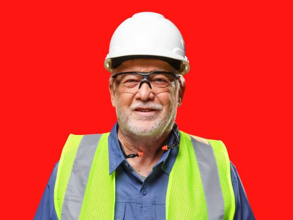 Man with white scruff wearing white hard hat, safety glasses, hi-visibility vest, and earplugs around his neck smiles at camera.