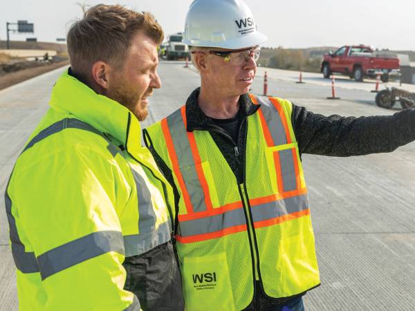Two men wearing high visibility clothing and one wearing a WSI hard hat at road construction site.