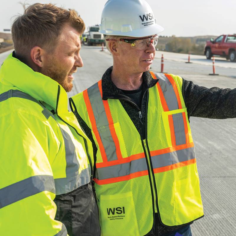 Two men wearing high visibility clothing and one wearing a WSI hard hat at road construction site.