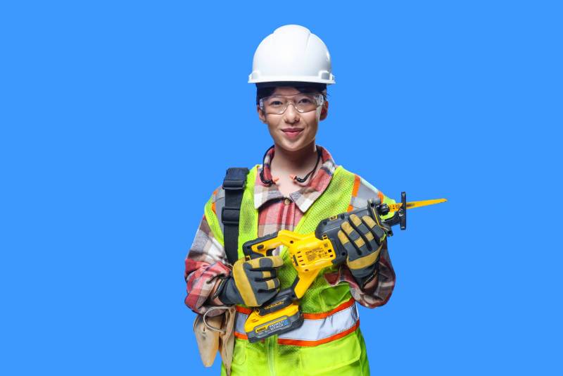 Woman wearing hard hat, safety glasses, hi-visibility vest, and safety gloves holding a power tool smiling at camera.