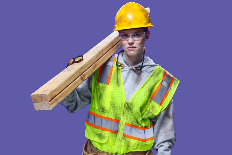 Woman wearing hard hat, safety glasses, gloves, hi-vis vest and holding two 2x4s over her shoulder.