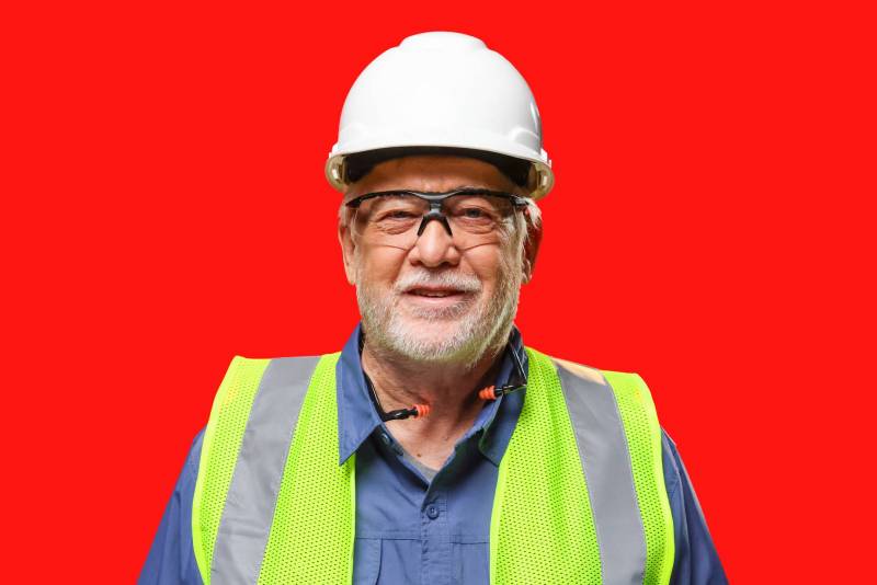 Man with white scruff wearing white hard hat, safety glasses, hi-visibility vest, and earplugs around his neck smiles at camera.